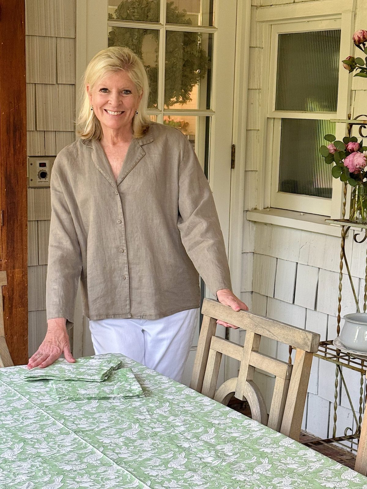 A woman with blonde hair stands next to a wooden chair and a table covered with a green and white patterned cloth on a porch.