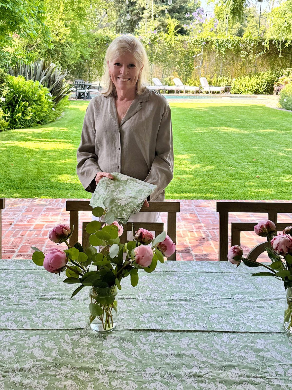 A woman stands behind an outdoor table set with a floral tablecloth and pink flowers, holding a matching napkin, with a garden and lawn in the background.