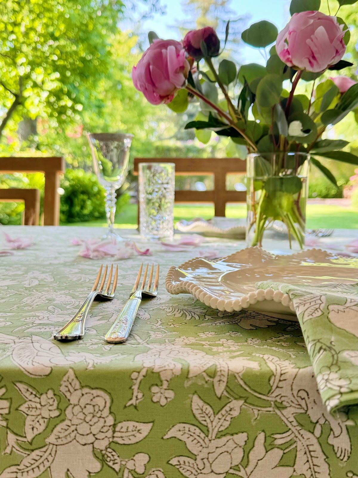 A close-up of a green floral tablecloth with two forks, plates, a glass, and a vase of pink flowers on an outdoor table. Trees and greenery are visible in the background.