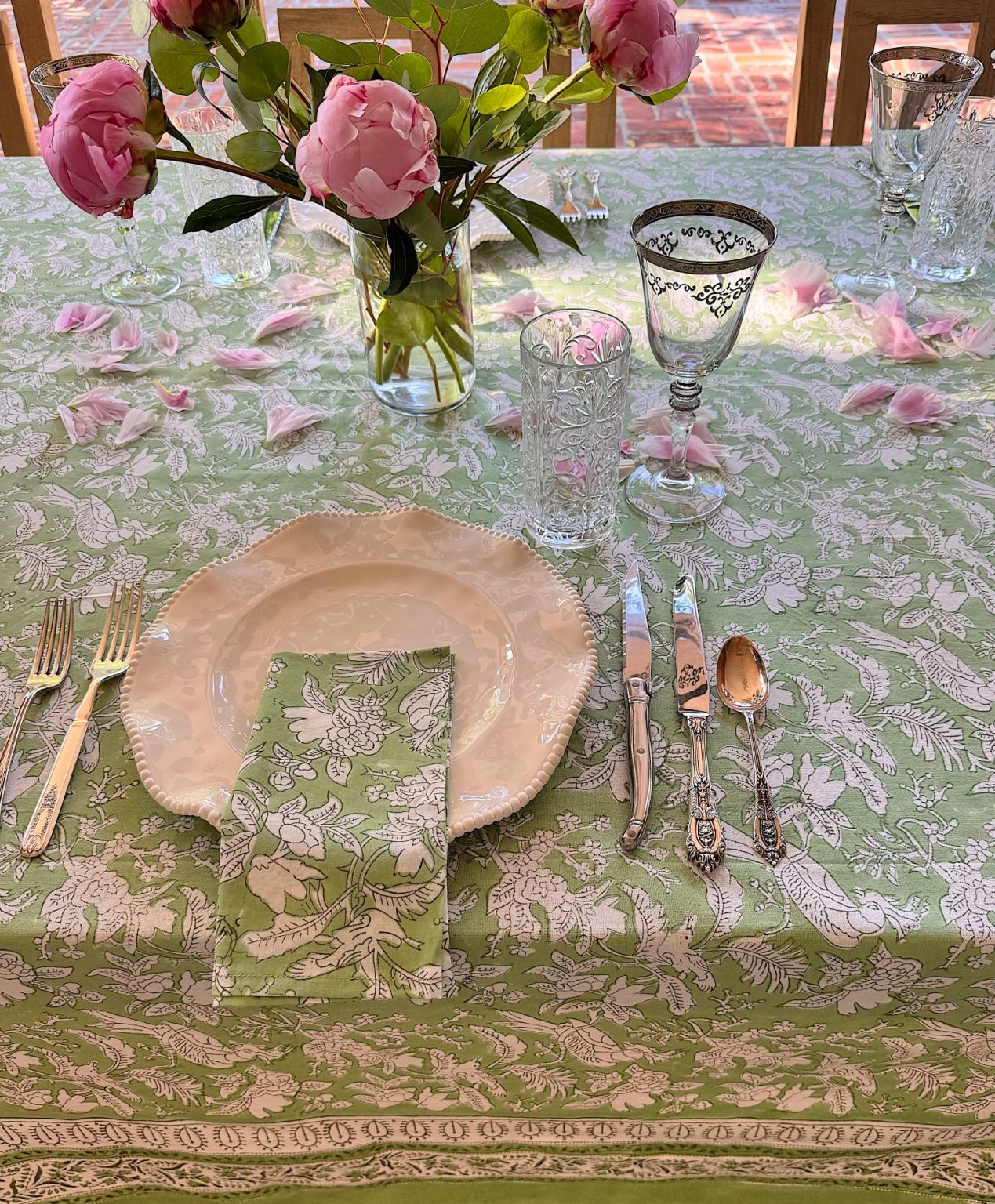 A table set with floral-patterned green tablecloth, matching napkin, white plate, glassware, cutlery, and a vase of pink peonies.