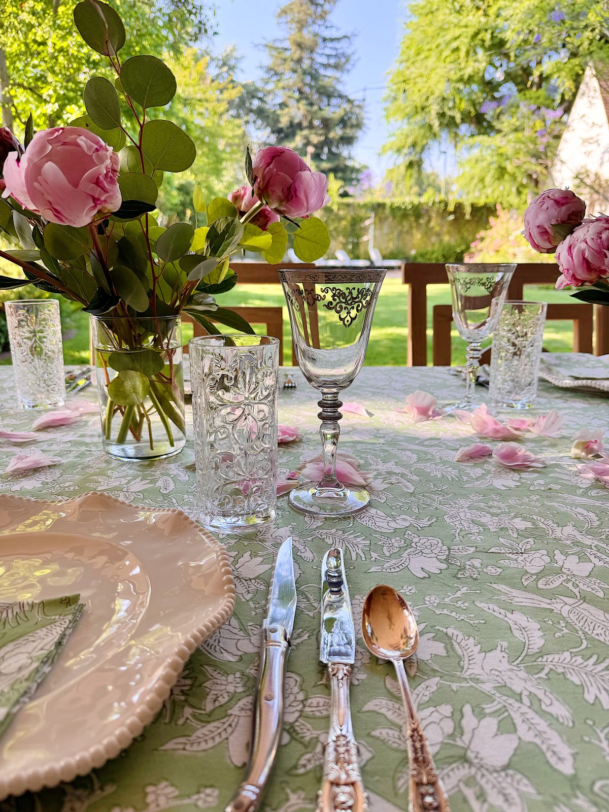A table set outdoors with floral-patterned dishes, vintage cutlery, etched glasses, and vases of pink roses on a green tablecloth, with a garden visible in the background.