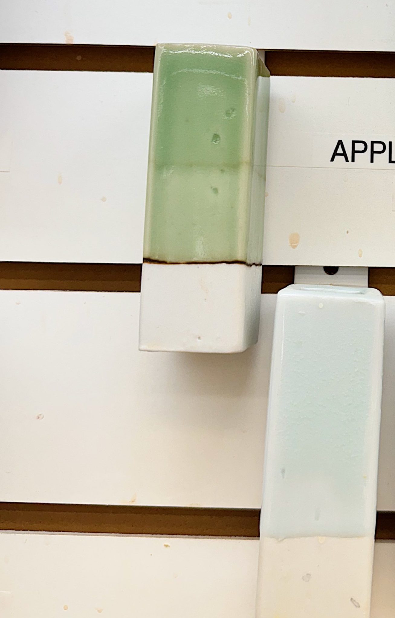 Rectangular ceramic tile with a glossy light green glaze on the top half and white on the bottom half, displayed vertically on a slotted white wall.
