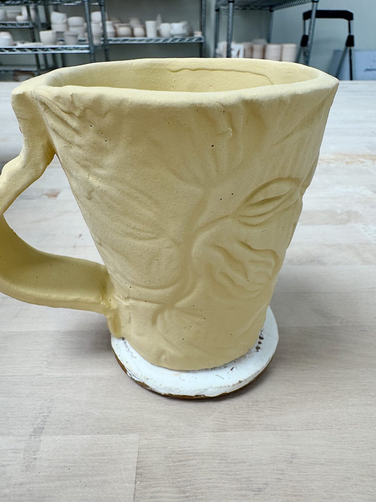A large, unfinished ceramic mug with a textured leaf or face design sits on a wooden table in a pottery studio.