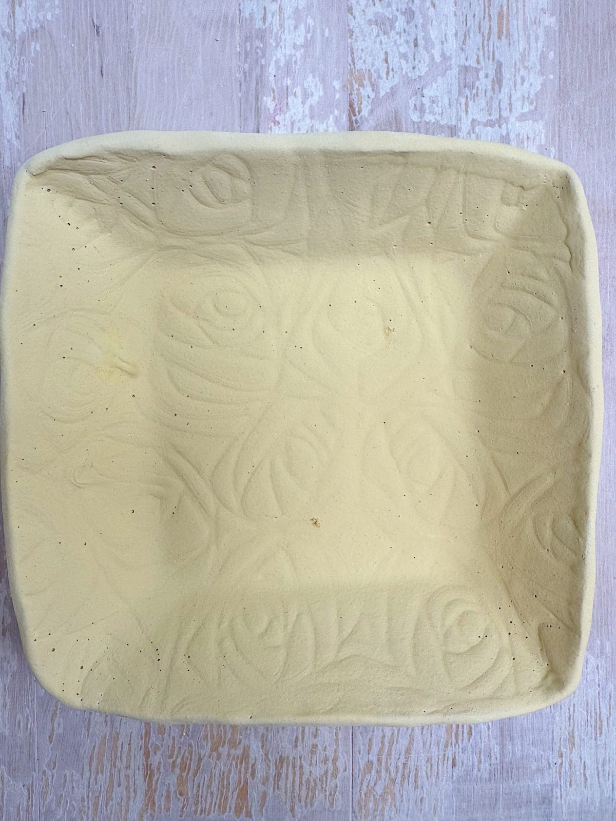 A square, shallow ceramic dish with a yellowish glaze and an embossed abstract swirl pattern, placed on a light wooden surface.