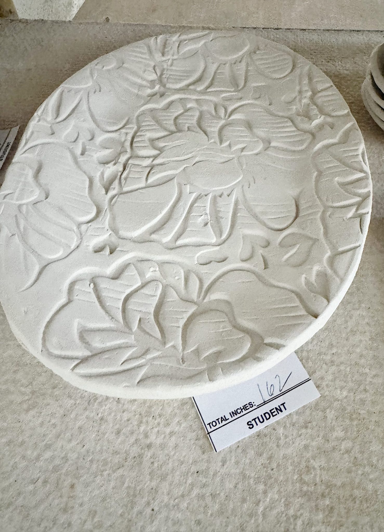 Round white clay plate with raised floral patterns rests on a table; a label beside it reads "TOTAL INCHES: 16 1/2 STUDENT.