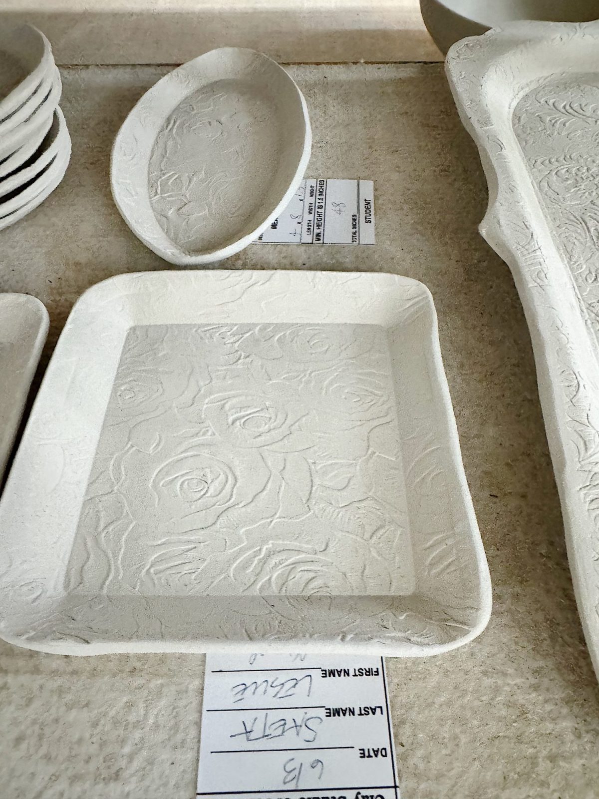 A white, square ceramic plate with a floral embossed pattern is displayed on a table next to other dishware, with a name tag labeled "Leslie" partially visible underneath.