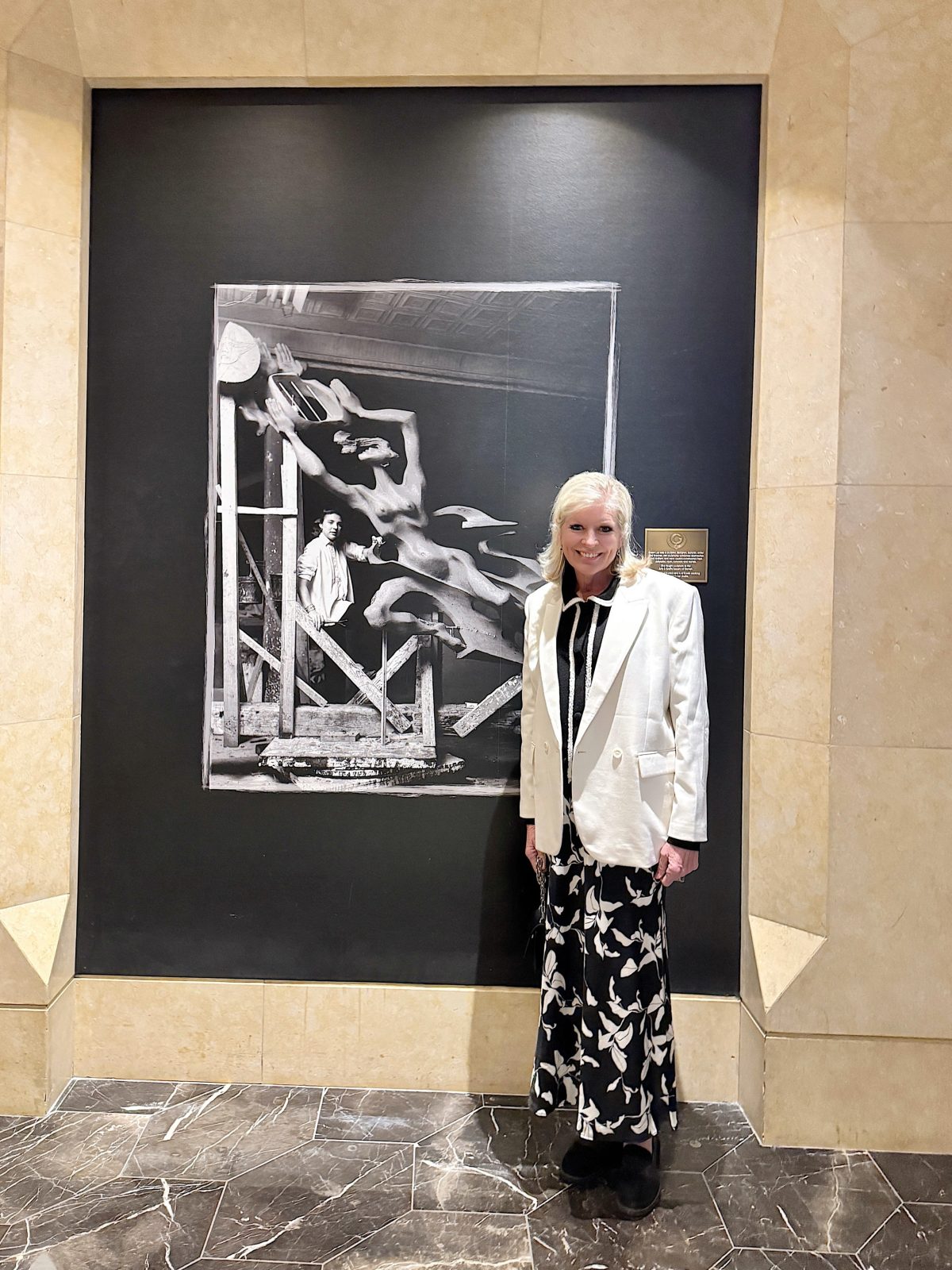 A woman in a white blazer and patterned skirt stands in front of a black-and-white artwork mounted on a stone wall.