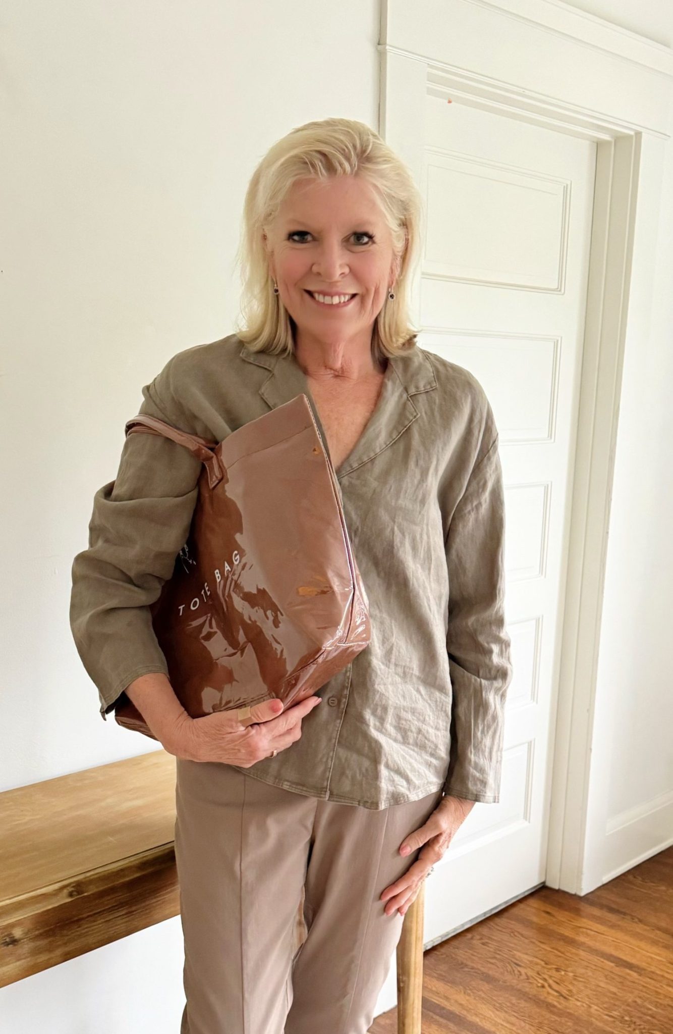 A woman with light hair stands indoors, smiling, holding a large brown tote bag. She wears a beige top and pants. A wooden table and white door are in the background.