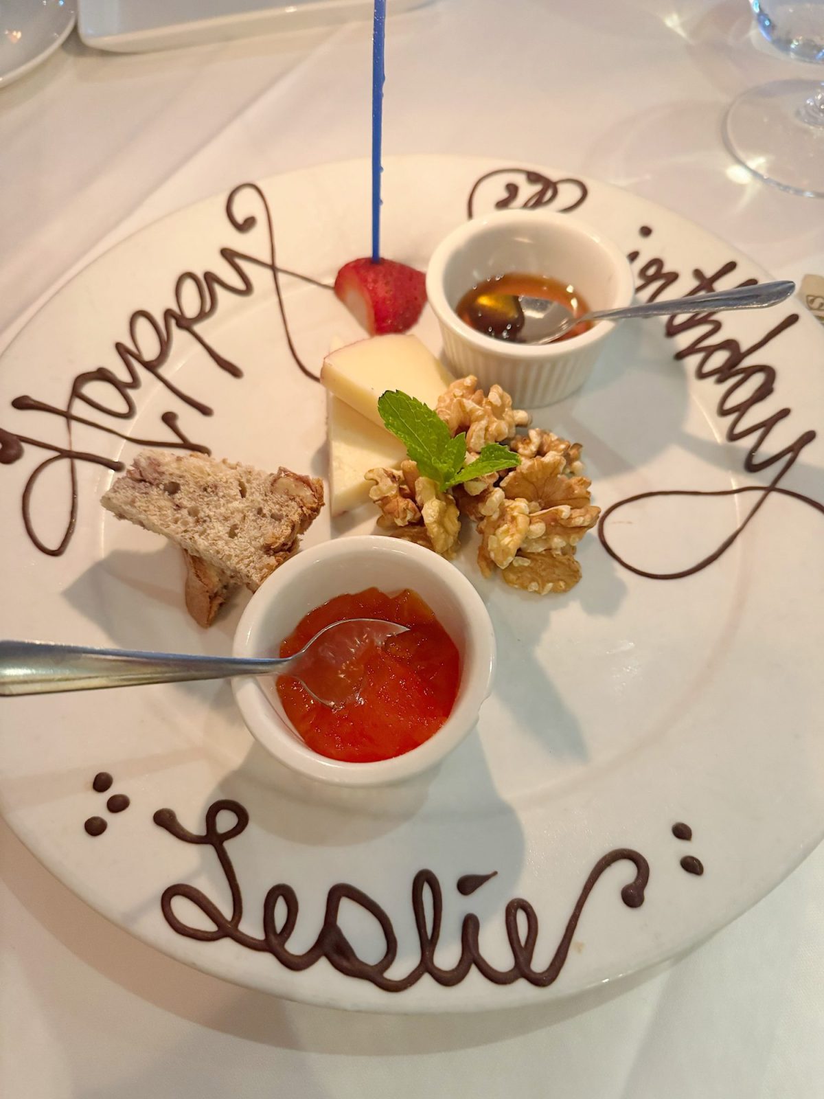 A dessert plate with “Happy Birthday Leslie” written in chocolate, holding walnuts, cheese, bread, strawberry, jam, and a sauce, with a lit blue candle.