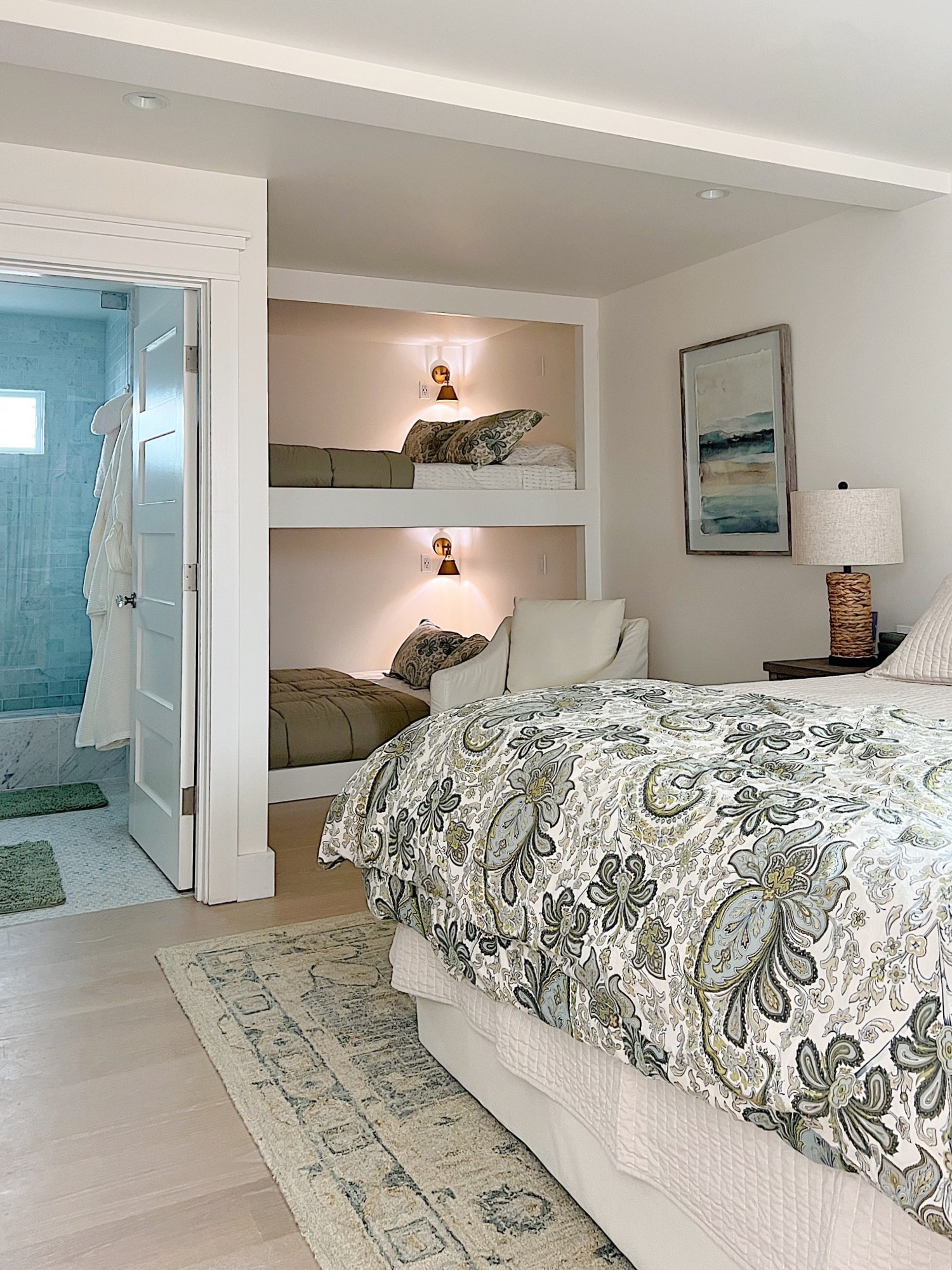 A bedroom with a patterned bedspread, bunk beds built into the wall, a bedside table with a lamp, and an open door leading to a bathroom.