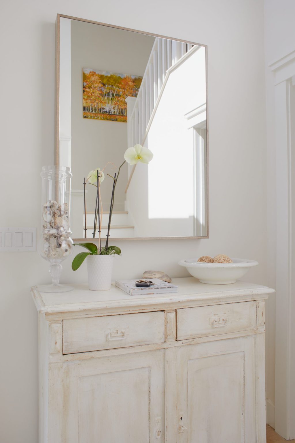 A white cabinet with drawers holds a potted orchid, a glass vase of shells, and a shallow bowl; a large mirror and wall art hang above, reflecting a staircase.