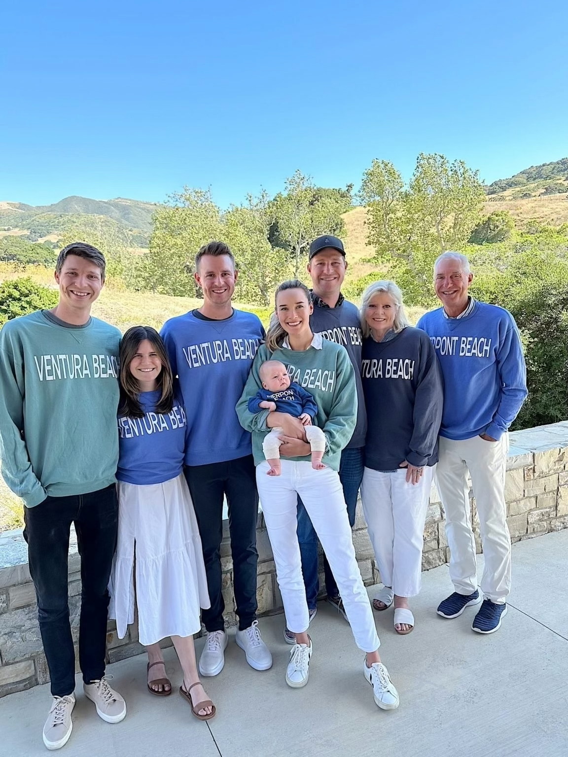 A group of seven adults and one baby stand outdoors, all wearing Ventura Beach sweatshirts, posing together in front of a scenic landscape during their summer travel.