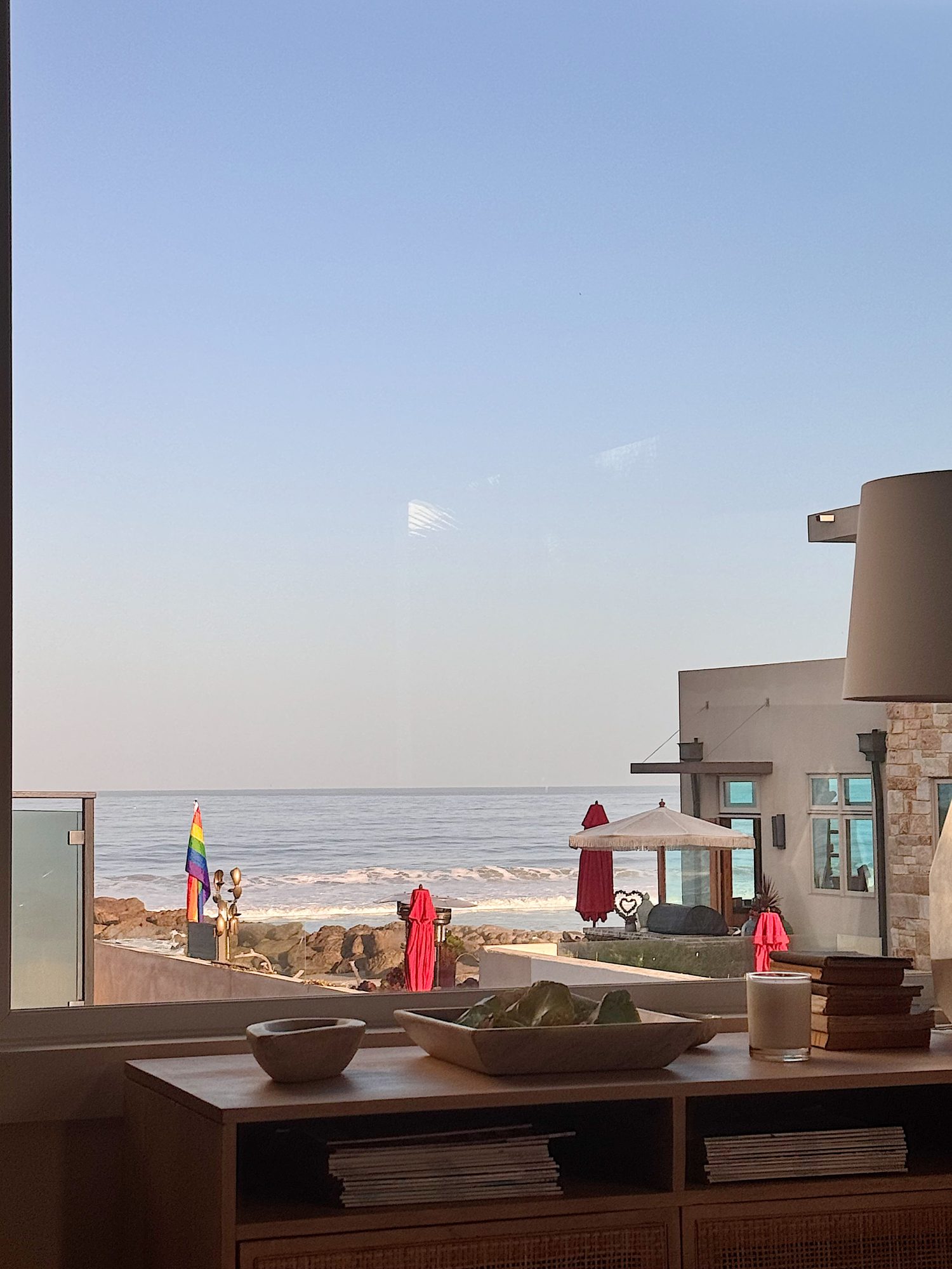 View through a window of a beachside scene with red umbrellas, a rainbow flag, lounge chairs, and calm ocean waves under a clear sky. Indoor furniture is visible in the foreground.