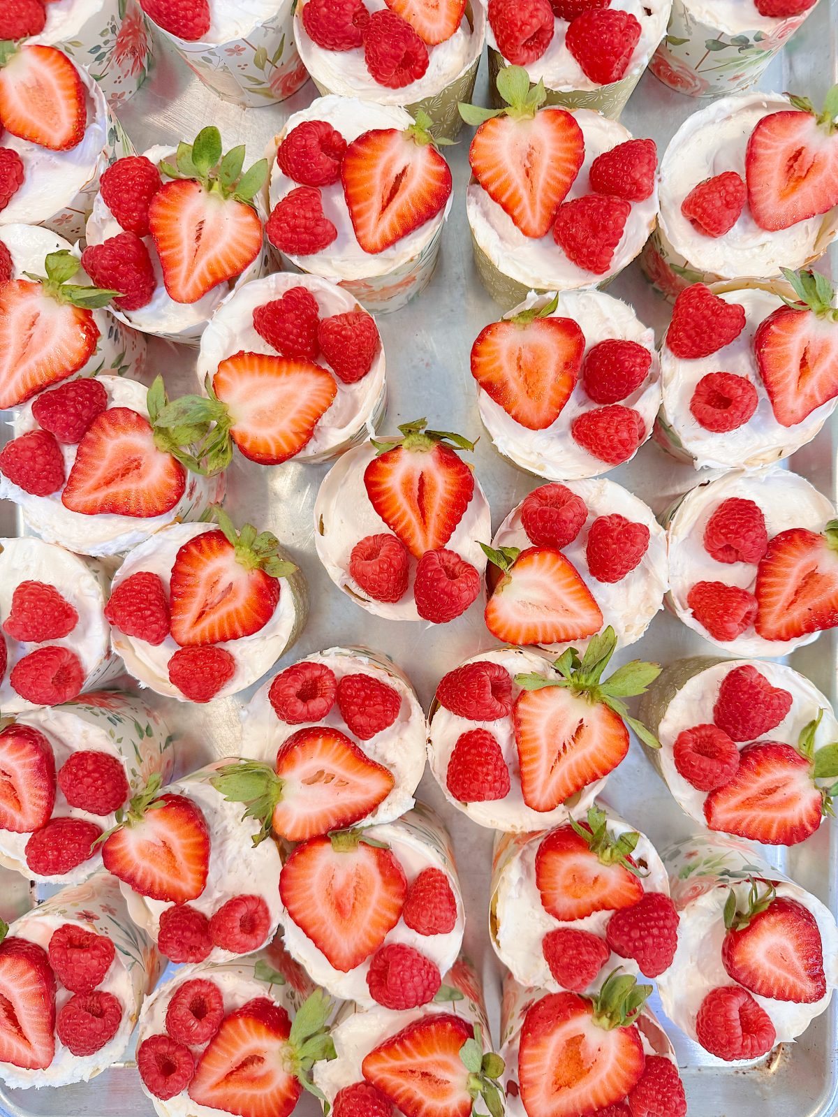 Rows of small desserts topped with whipped cream, fresh strawberries, and raspberries, arranged closely together on a tray.