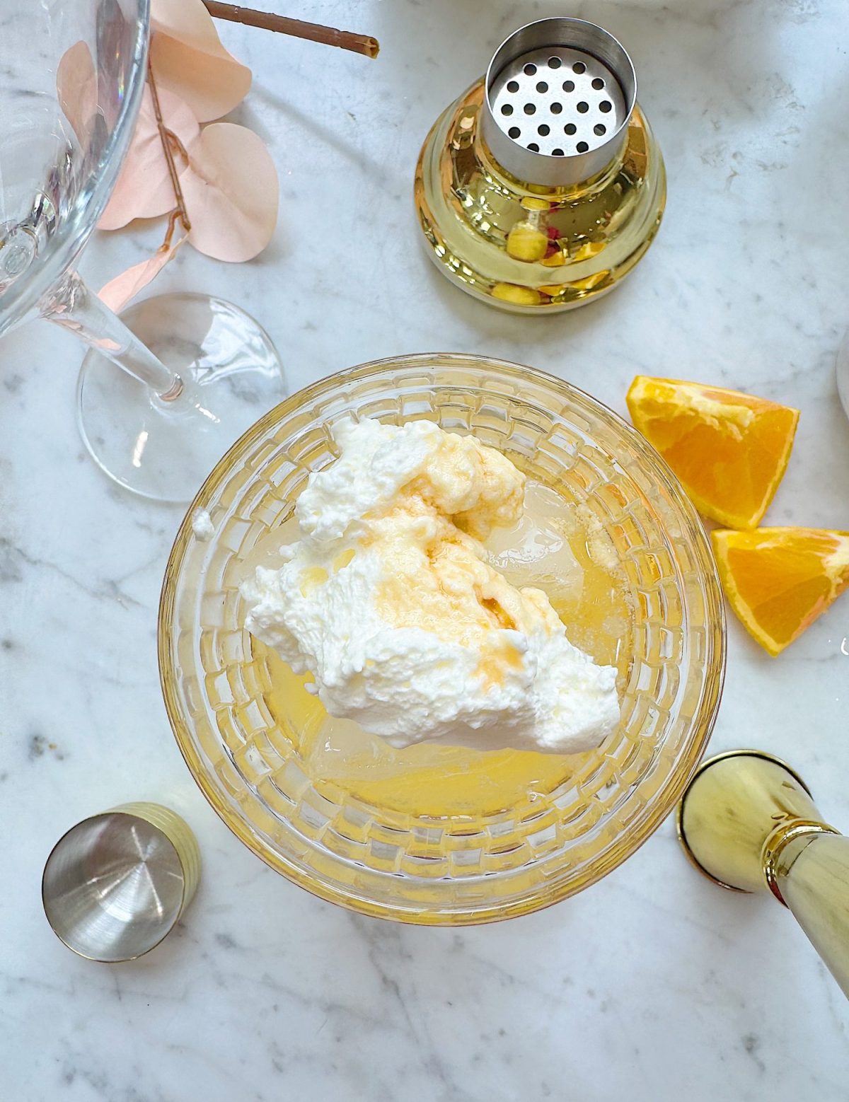 A glass bowl with a creamy white topping over a yellow drink, surrounded by orange wedges, a jigger, cocktail shaker, and a wine glass on a marble surface.