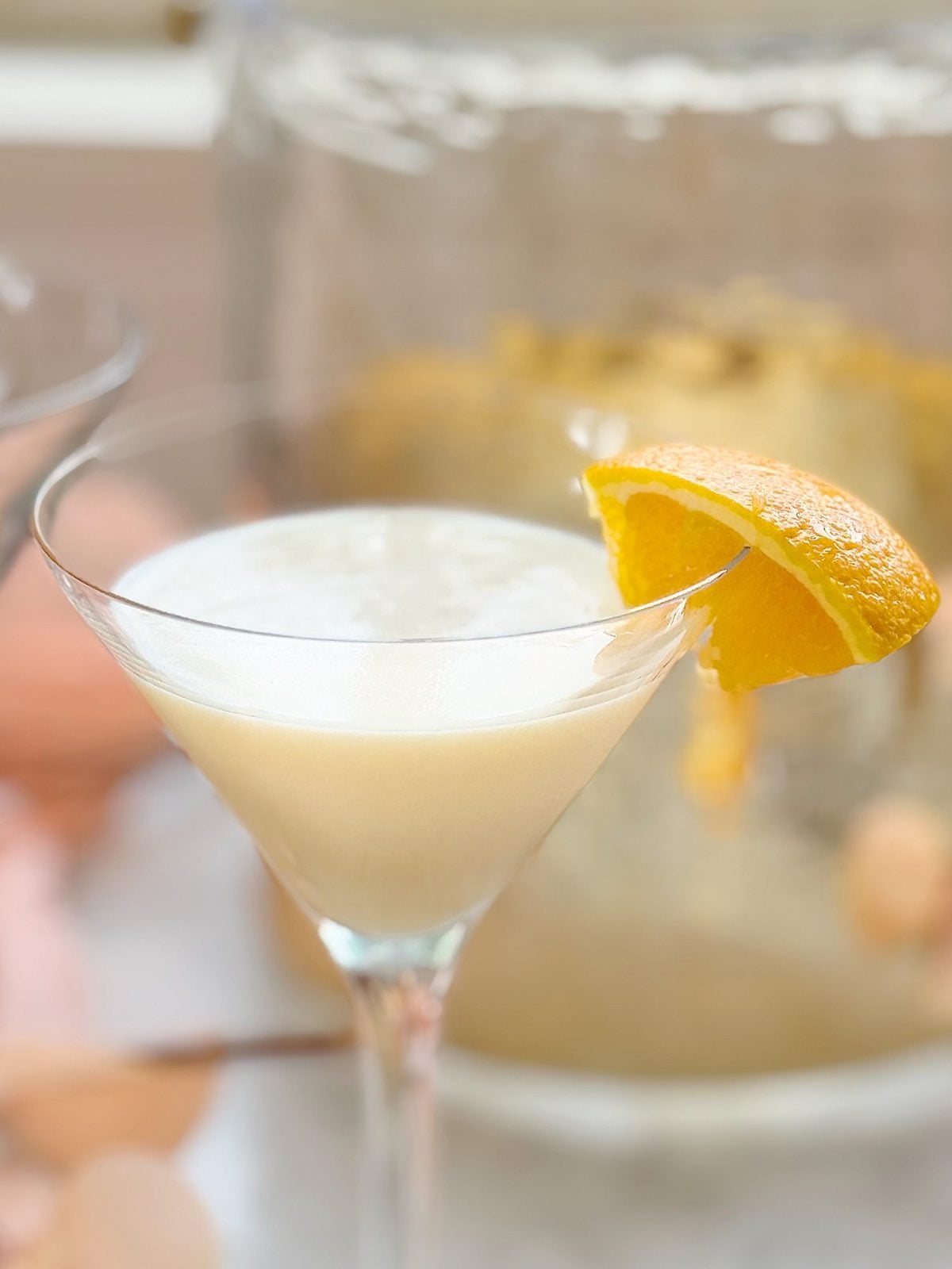 A martini glass filled with a pale, frothy cocktail garnished with a lemon wedge on the rim.