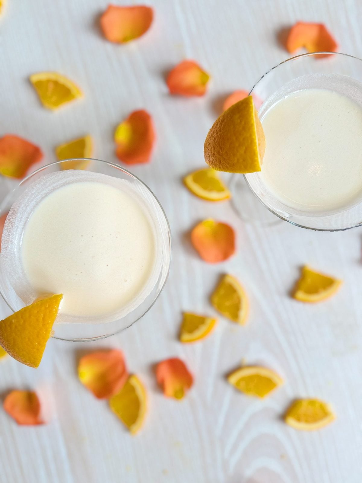Two cocktails garnished with orange wedges on a white surface, surrounded by scattered orange slices and pink rose petals.