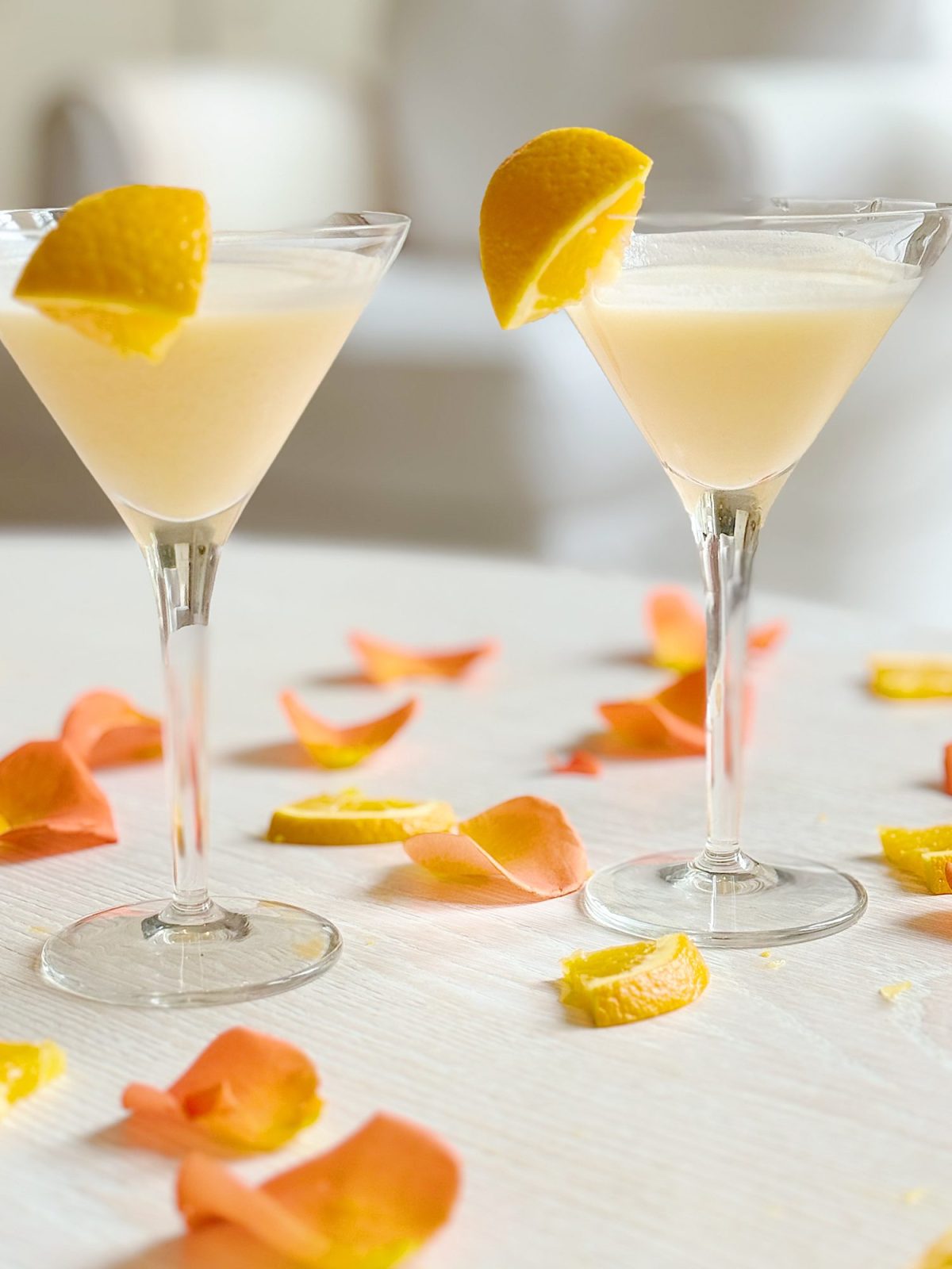 Two martini glasses filled with a pale yellow drink, each garnished with an orange slice, sit on a white surface scattered with orange rose petals and orange slices.