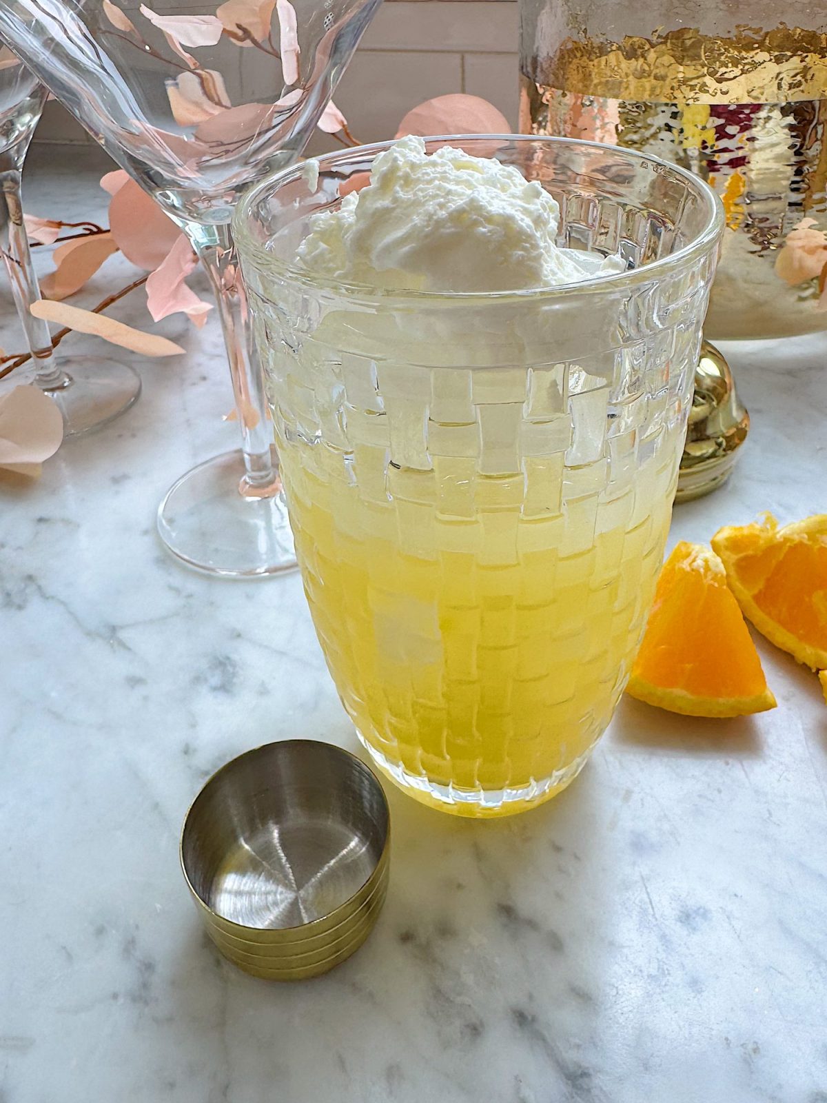 A glass of yellow cocktail with a dollop of whipped cream on top sits on a marble surface, surrounded by orange slices, a metal jigger, and cocktail glasses.