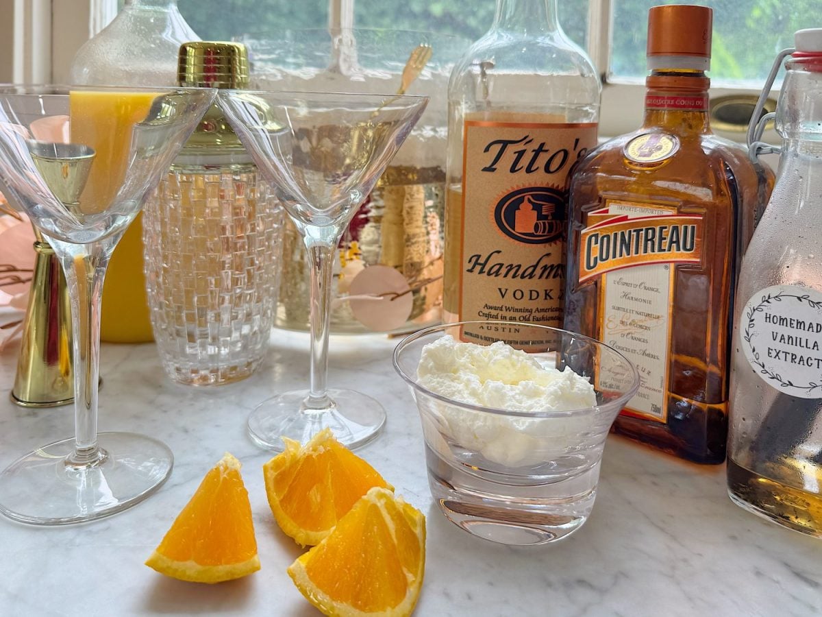 A marble countertop displays martini glasses, orange wedges, a bowl of whipped cream, vodka, Cointreau, homemade vanilla extract, a cocktail shaker, and a jug of orange juice.