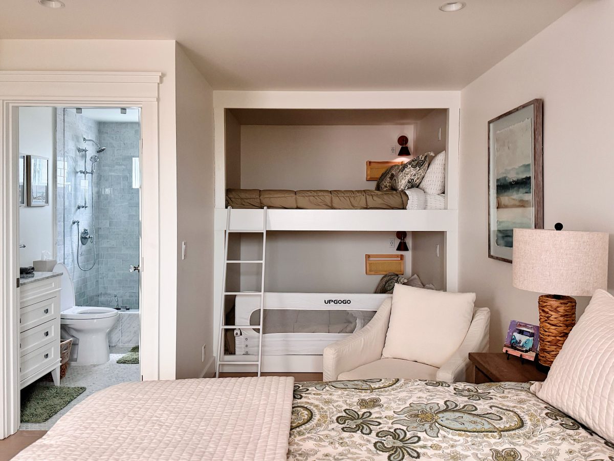 A bedroom with a patterned bed, an armchair, a nightstand with a lamp, bunk beds, and an en suite bathroom visible in the background.