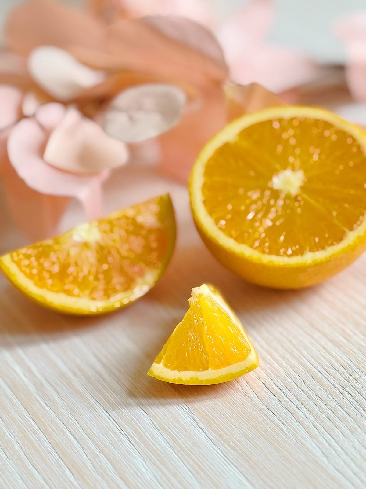 A halved orange and two orange wedges are placed on a light wooden surface with soft pink flowers in the background.
