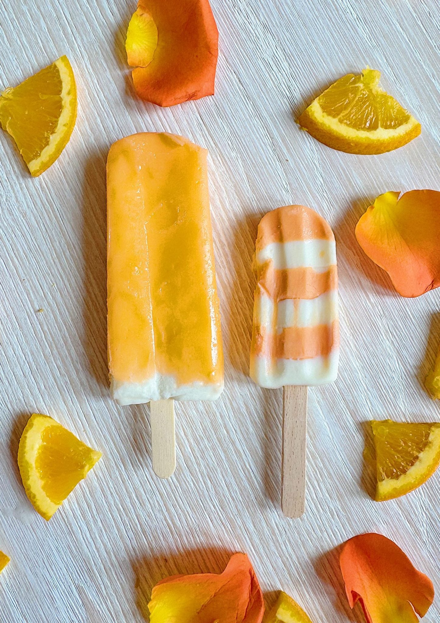 Two orange popsicles, one solid and one striped with white, are placed on a light wood surface surrounded by orange slices and orange flower petals.