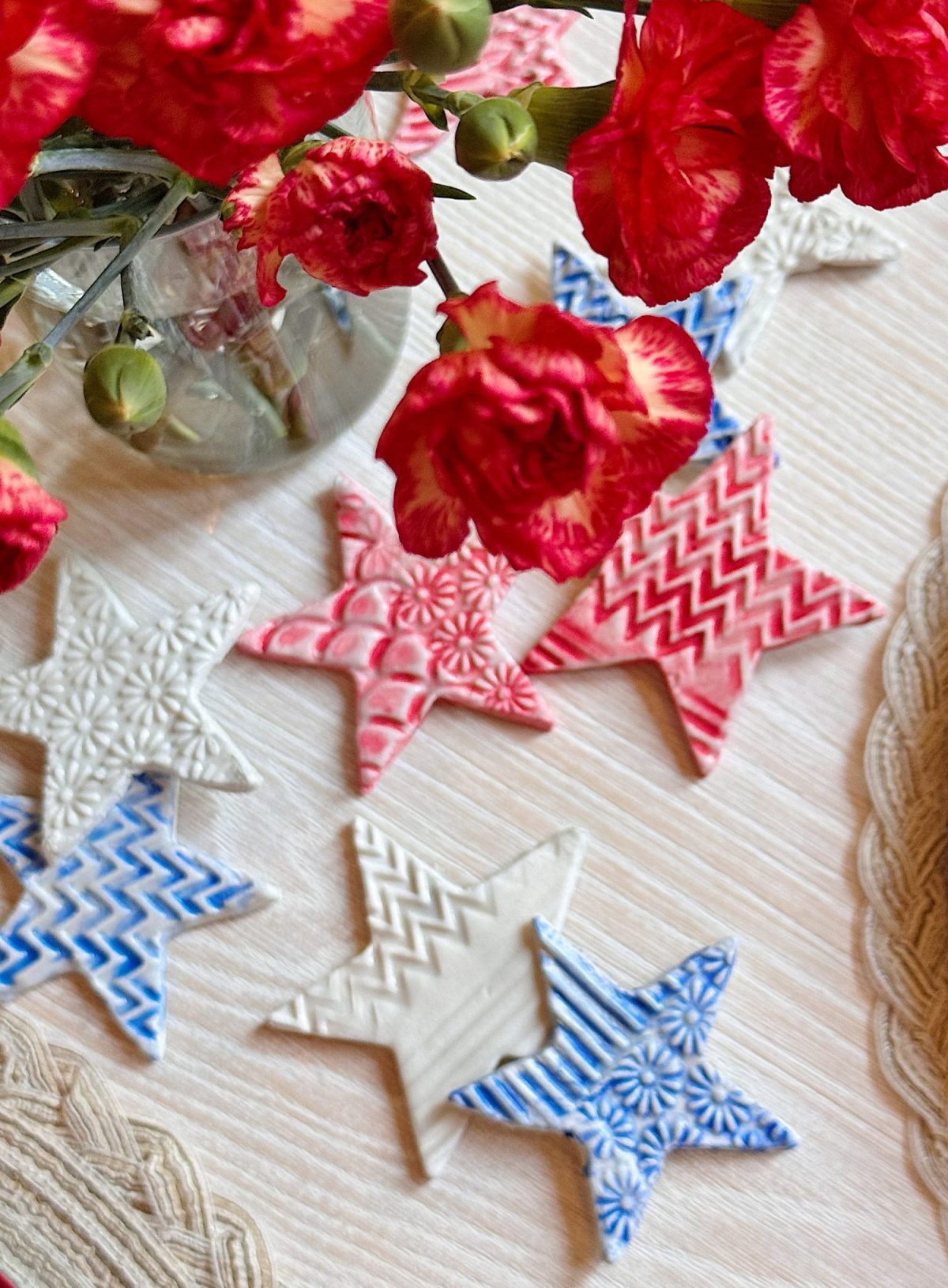 Red and pink flowers in a vase next to star-shaped ceramic ornaments in red, blue, and white with various patterns, arranged on a light wooden table.