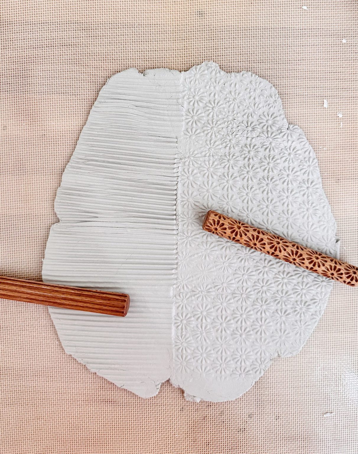 A sheet of clay is half imprinted with parallel lines and half with a floral pattern, using two textured wooden rolling pins lying on top.