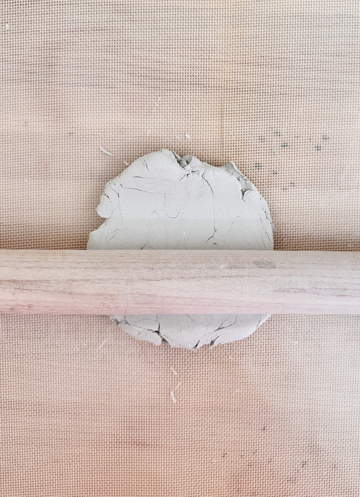 A ball of gray clay being flattened with a wooden rolling pin on a textured surface.