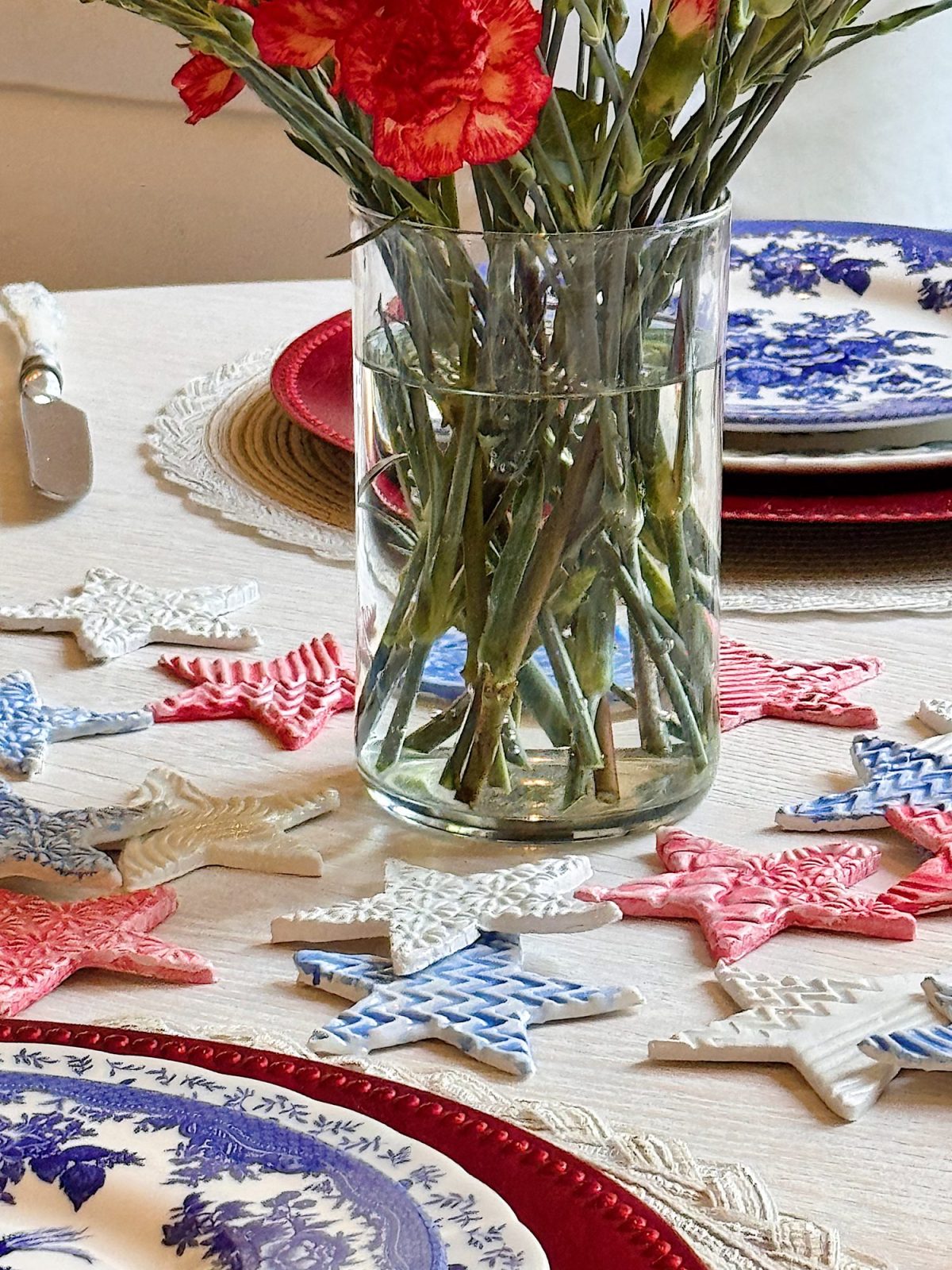 A glass vase with red flowers is on a table set with blue and white plates and surrounded by red, white, and blue star-shaped decorations.