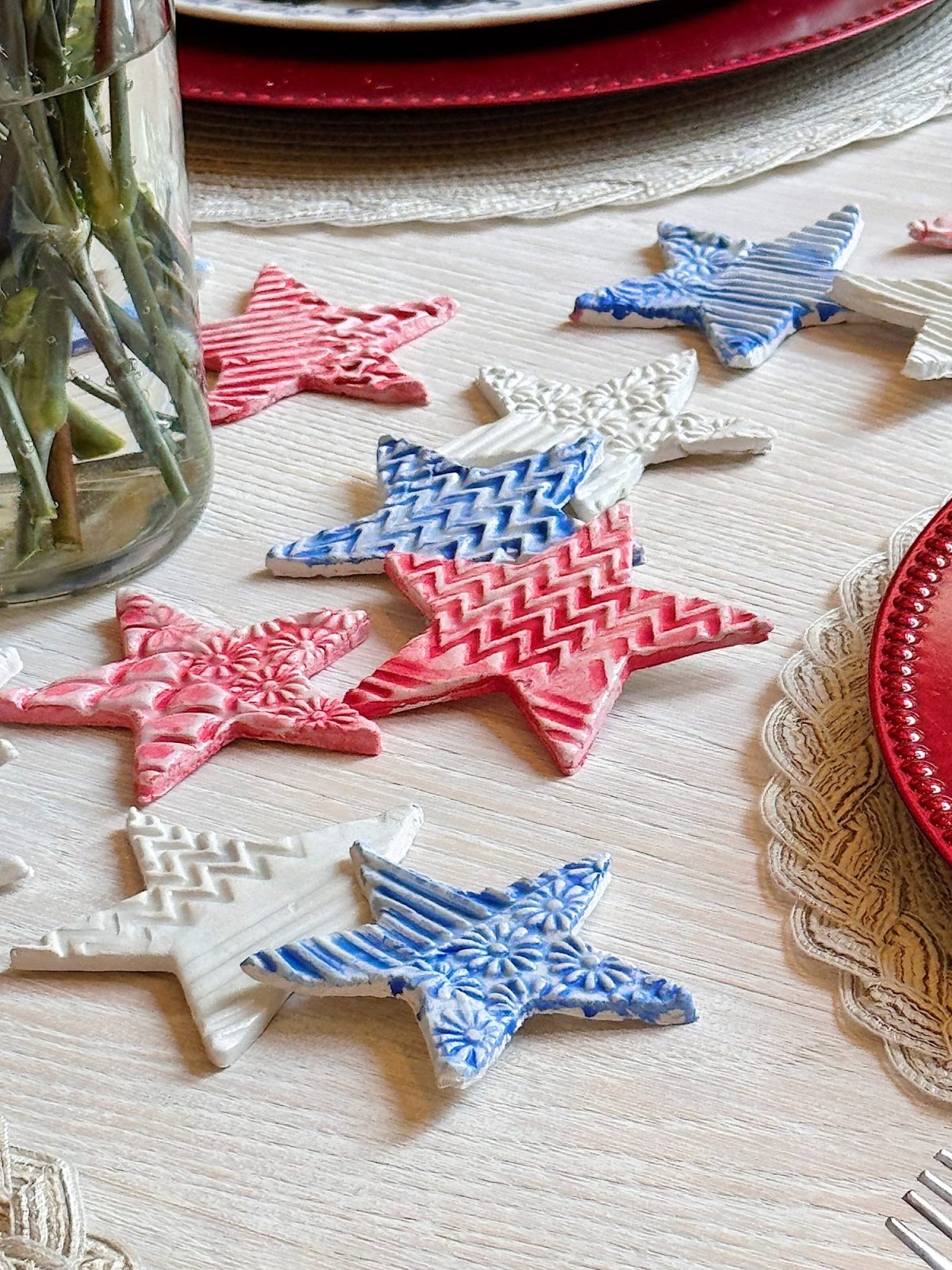 Red, white, and blue star-shaped ornaments with textured patterns are arranged on a light wooden table next to a vase and lace placemats.