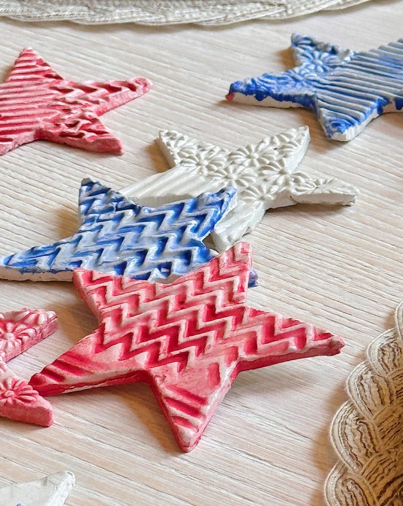 Red, white, and blue star-shaped clay ornaments with textured patterns are arranged on a light wooden surface.