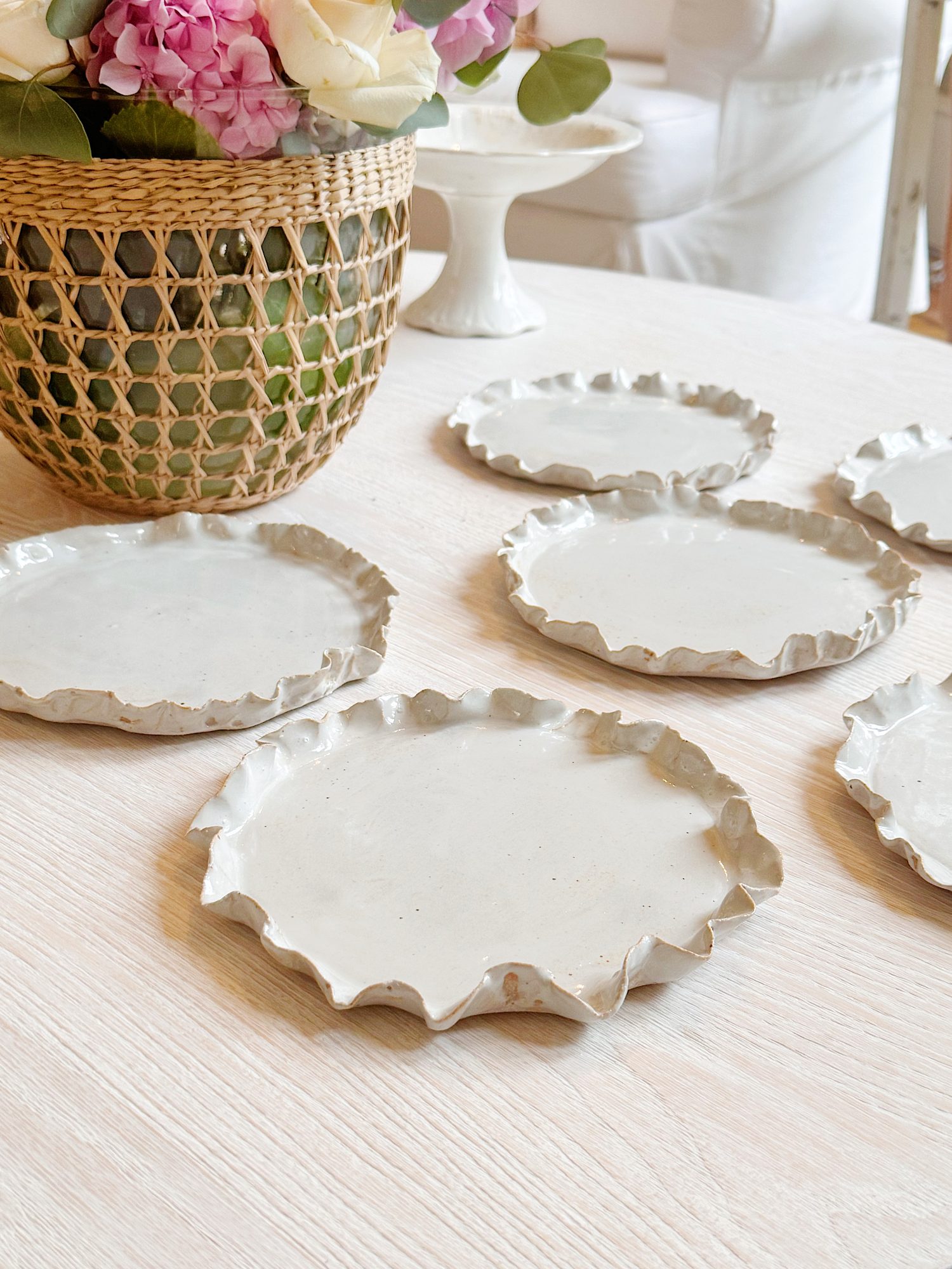 Six white ceramic plates with ruffled edges are arranged on a light wooden table near a woven basket with pink and white flowers.