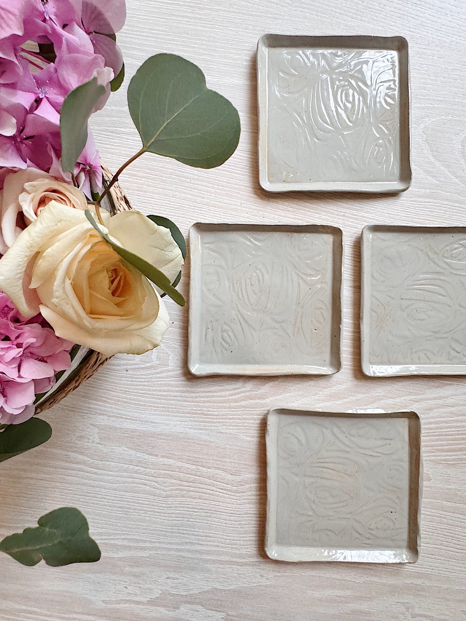 Four square ceramic plates with an embossed swirl pattern are arranged on a light wooden surface next to a bouquet of pink and cream flowers with green leaves.