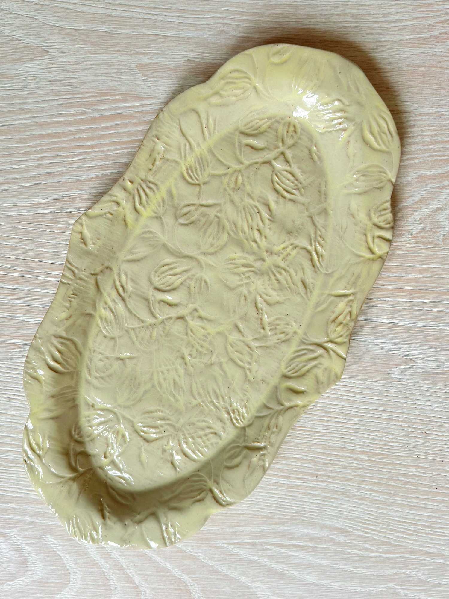 An oval-shaped ceramic platter with a scalloped edge and raised floral pattern, glazed in a light yellow color, placed on a light wood surface.