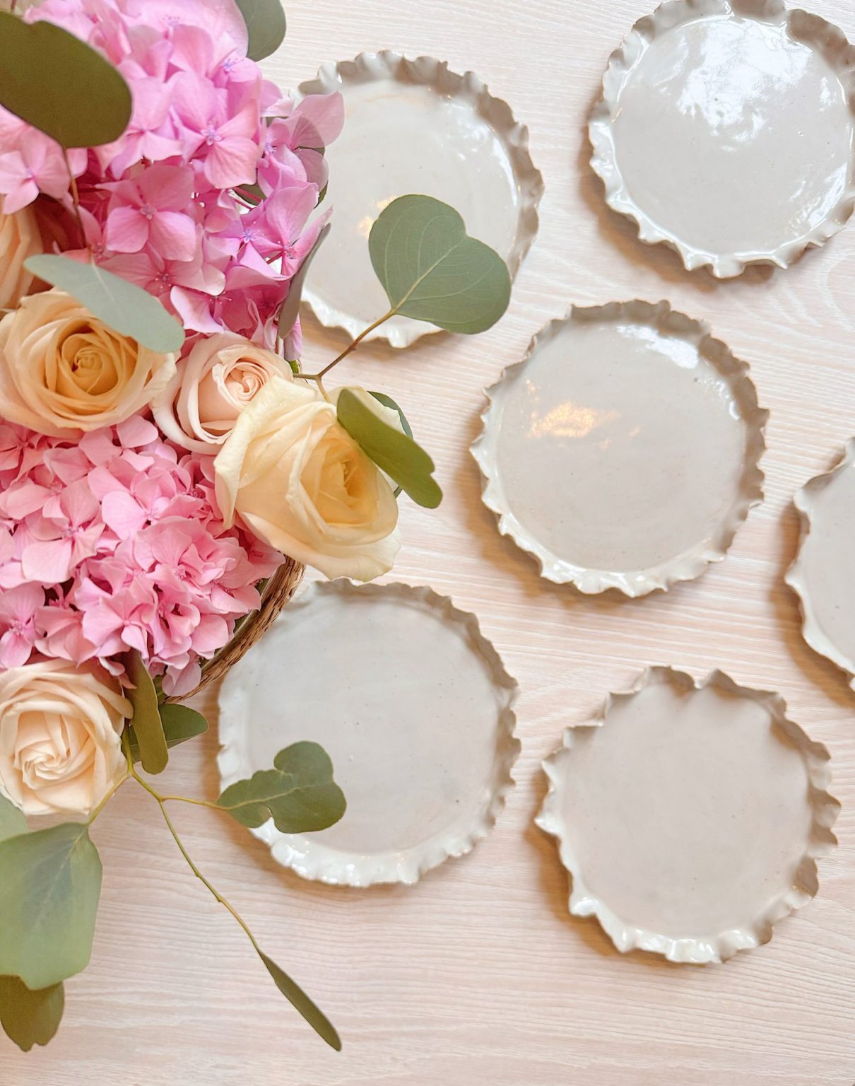 A bouquet of pink hydrangeas and cream roses next to seven round, fluted-edge ceramic plates on a light wooden surface.
