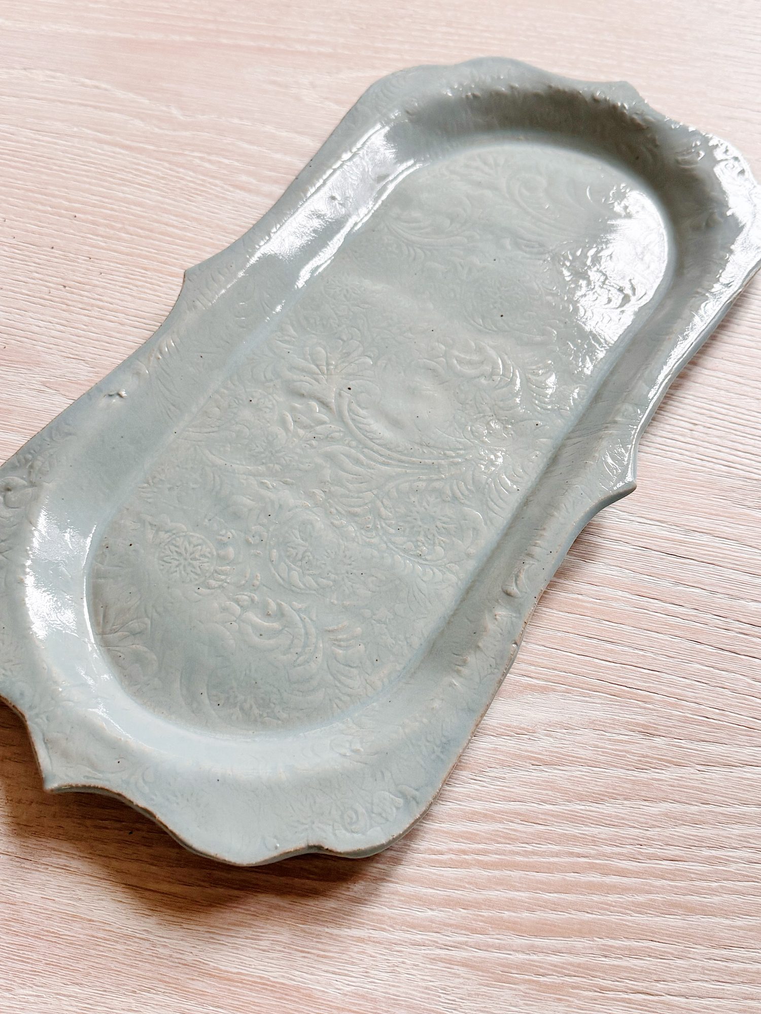 A rectangular, light gray ceramic tray with ornate embossed floral patterns, placed on a light wooden surface.