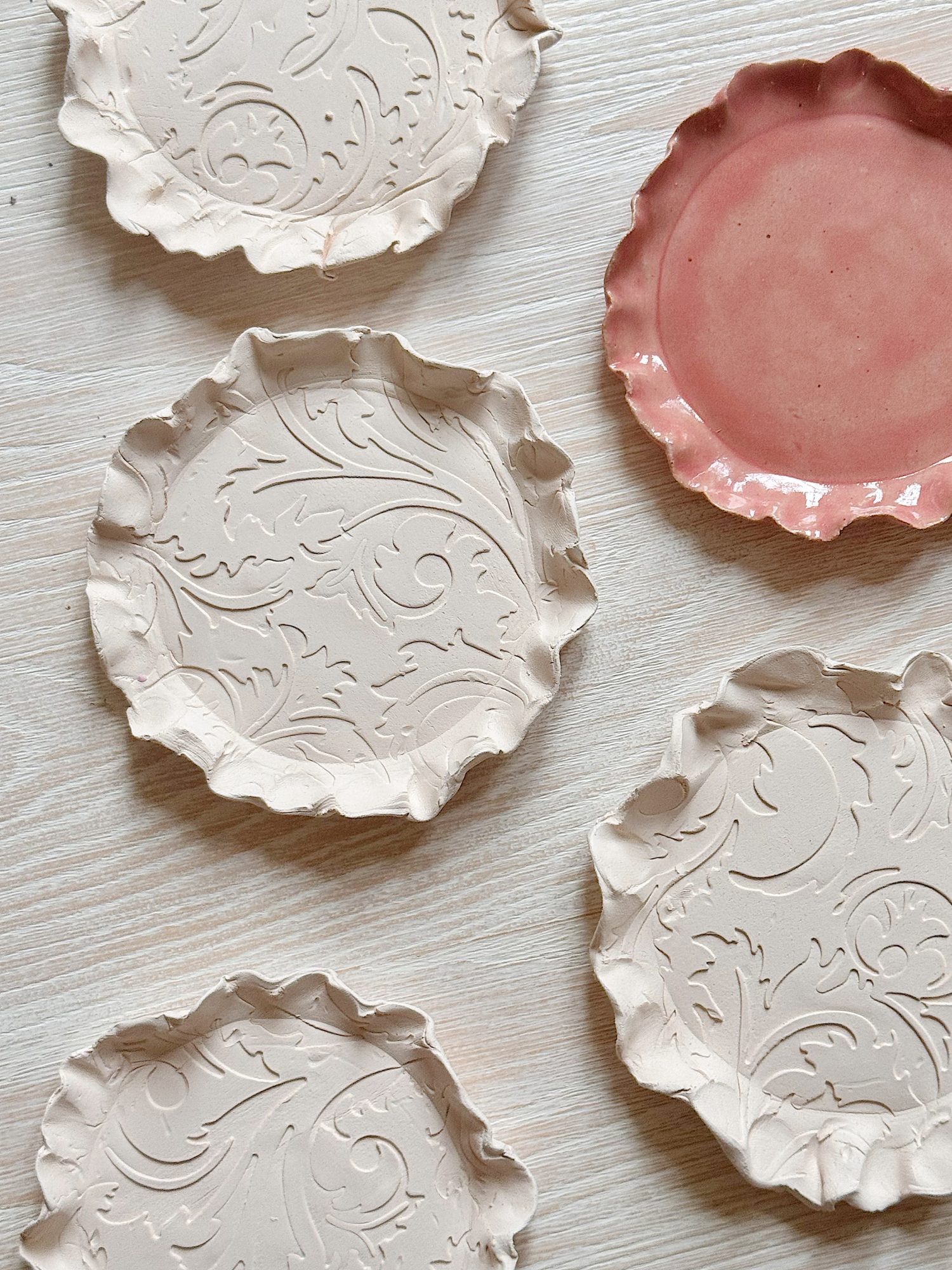 Five ceramic plates with wavy edges, four cream-colored with embossed swirls, and one solid pink, arranged on a light wooden surface.