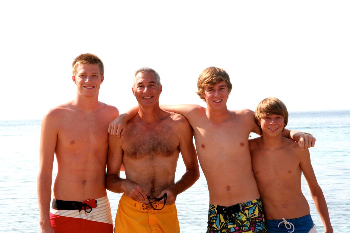 Four shirtless people in swim trunks stand side by side on a beach, smiling at the camera with the ocean in the background.