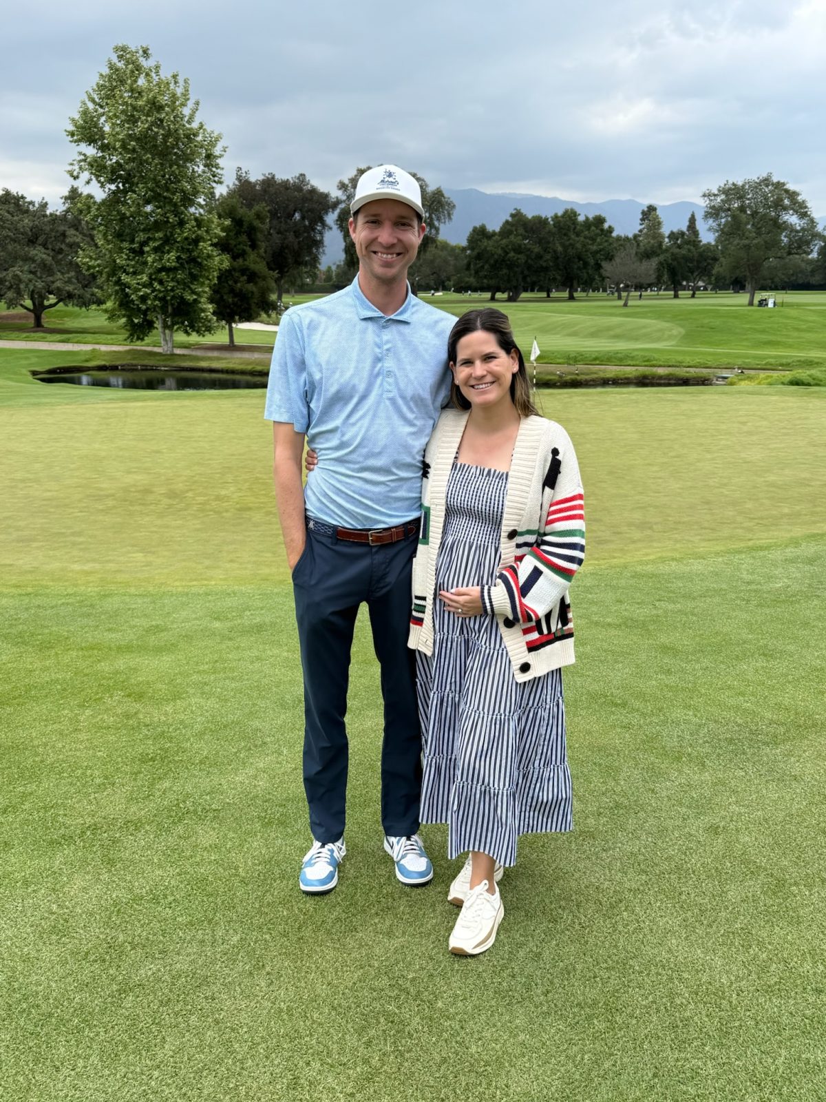 A man and a woman stand together on a golf course green, smiling at the camera. The man wears a golf outfit and cap; the woman wears a striped dress and cardigan.