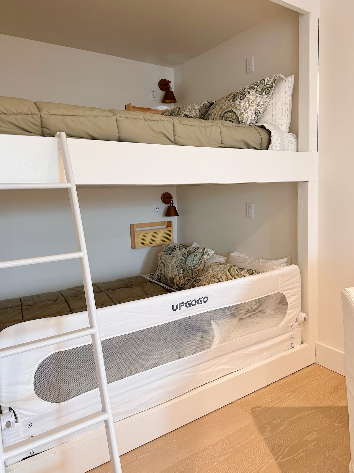 A white bunk bed with tan bedding and patterned pillows; the lower bunk has a white bed rail labeled "UPGOGO" and a ladder is positioned on the left side.