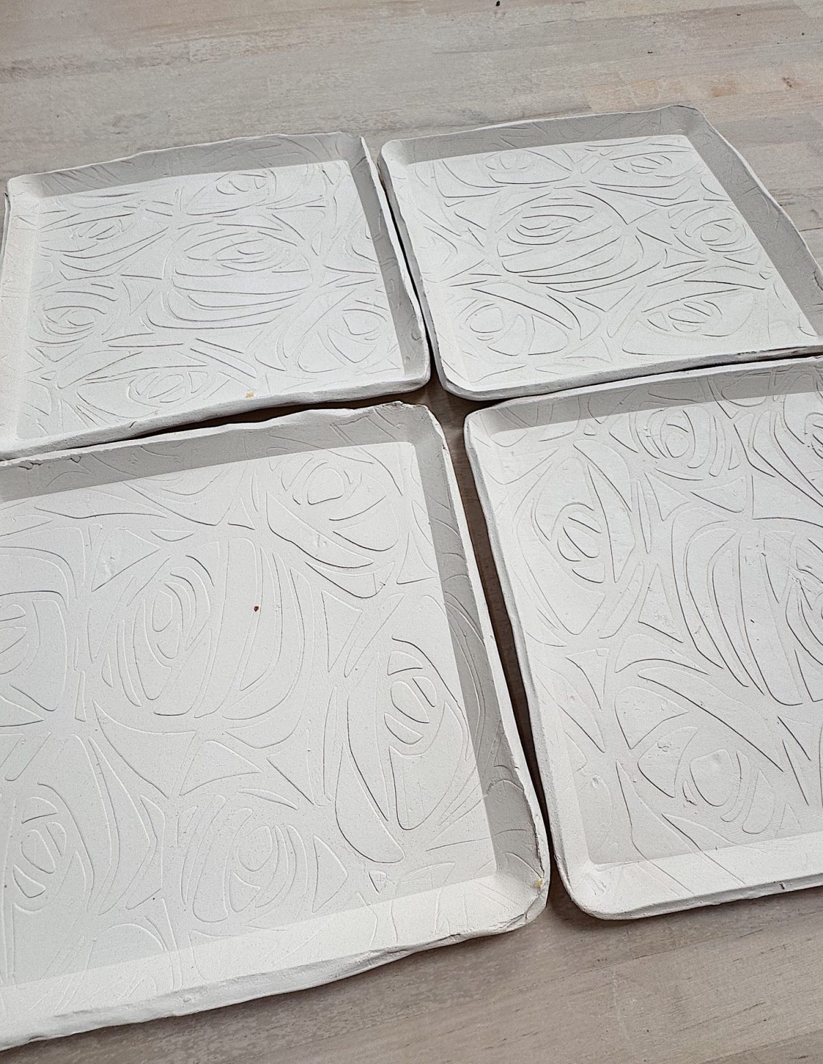 Four white, square ceramic plates with abstract, raised swirl patterns are arranged in a grid on a light wooden surface.