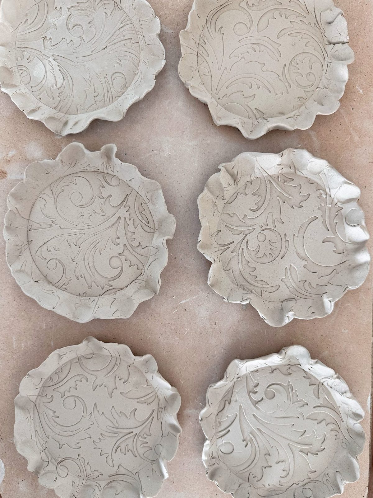 Six unglazed clay plates with embossed floral and leaf patterns and scalloped edges arranged on a flat surface.