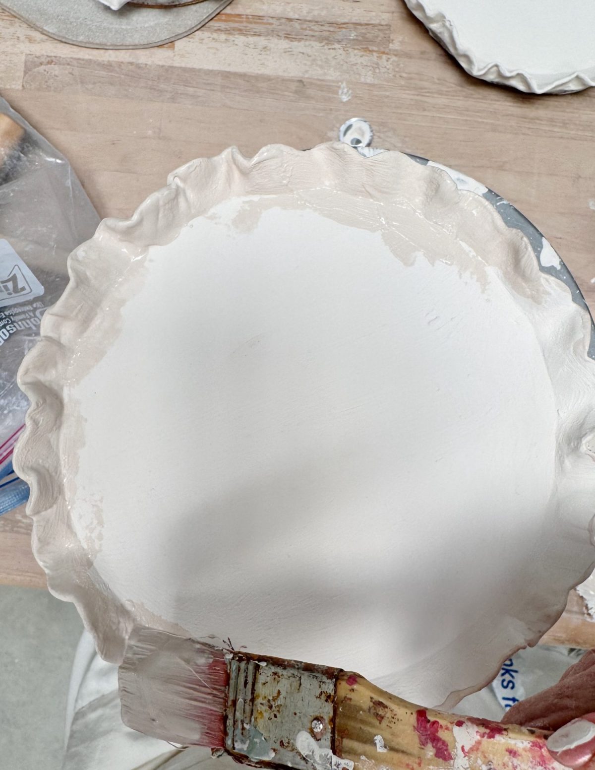 A hand holds a paintbrush applying white glaze to an unbaked ceramic plate with a ruffled edge on a wooden table.