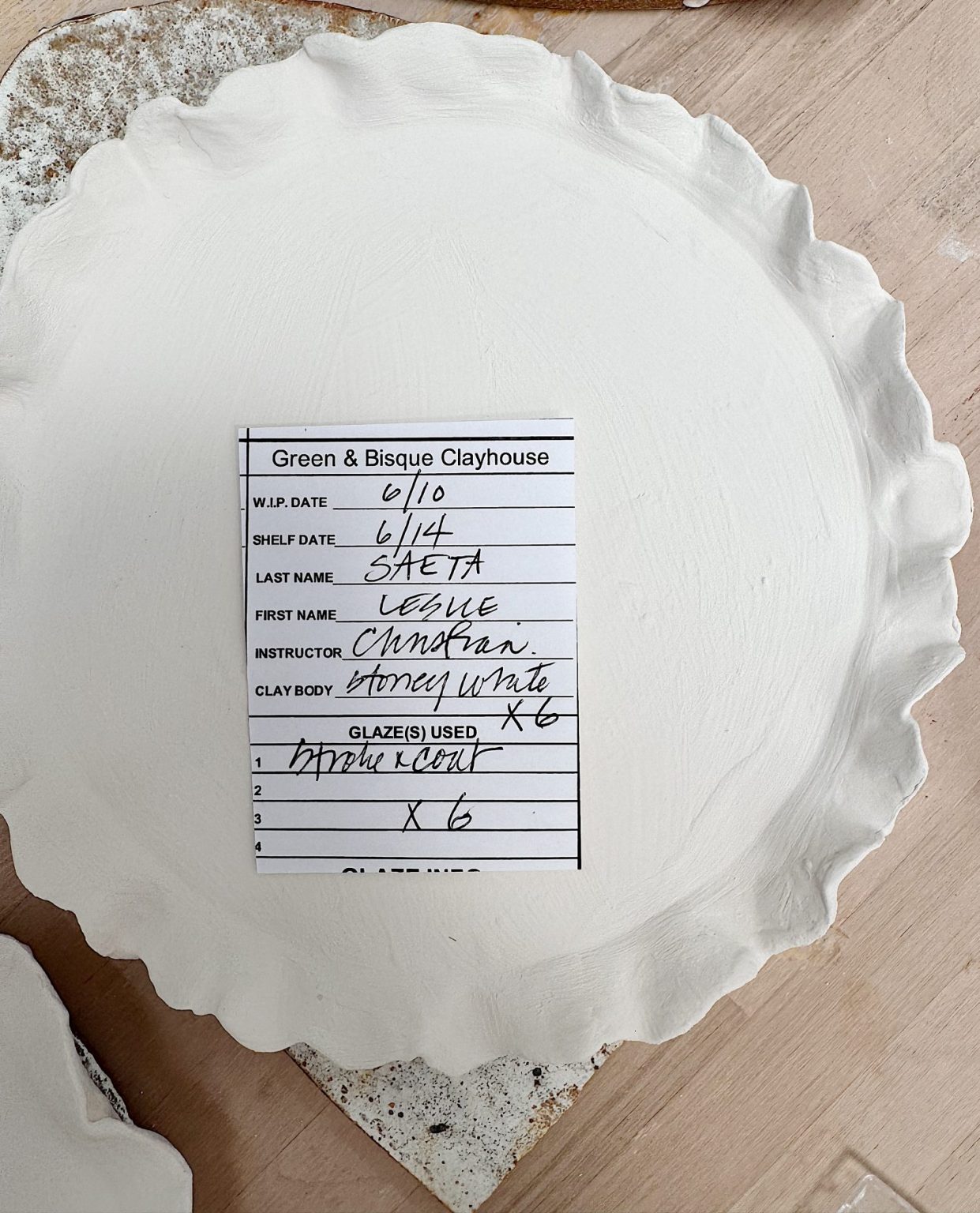 A round, unglazed white ceramic plate sits on a table with a workshop label listing details of the piece, including date, name, instructor, and materials used.