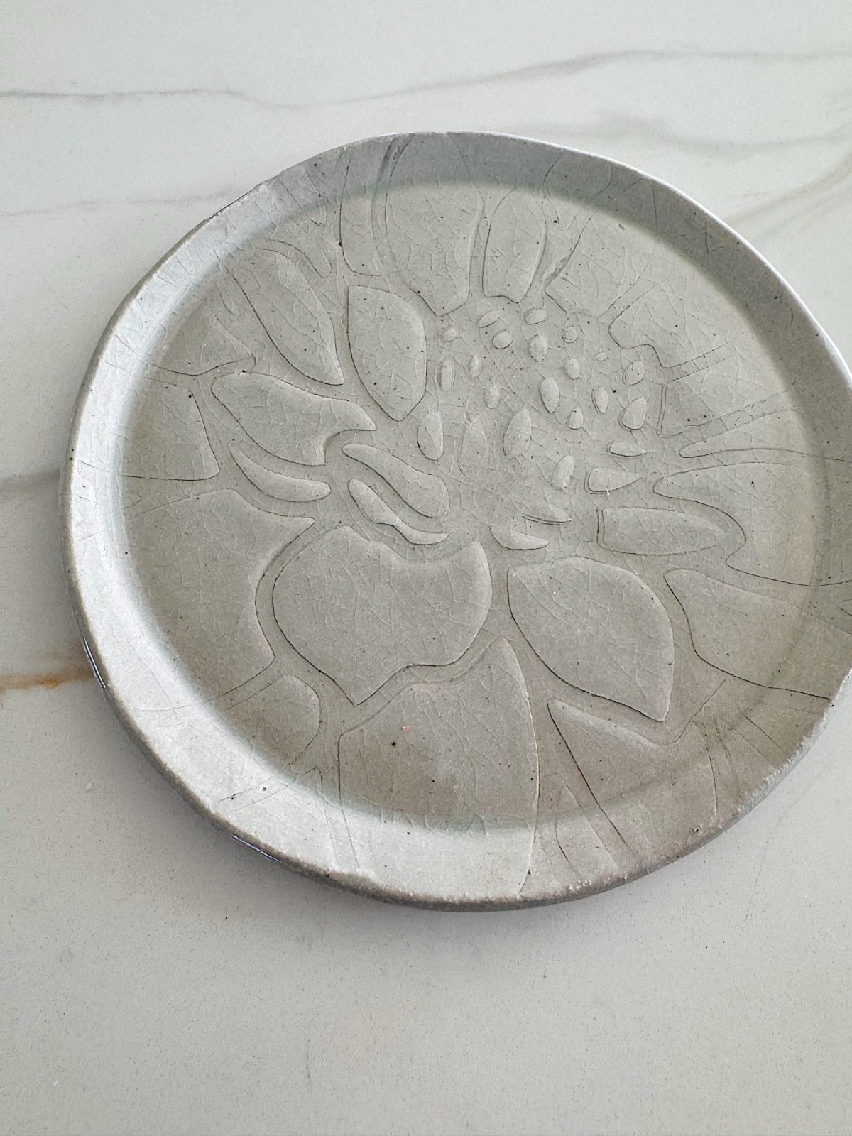 A round, light gray ceramic plate with an embossed floral pattern sits on a white marble surface.