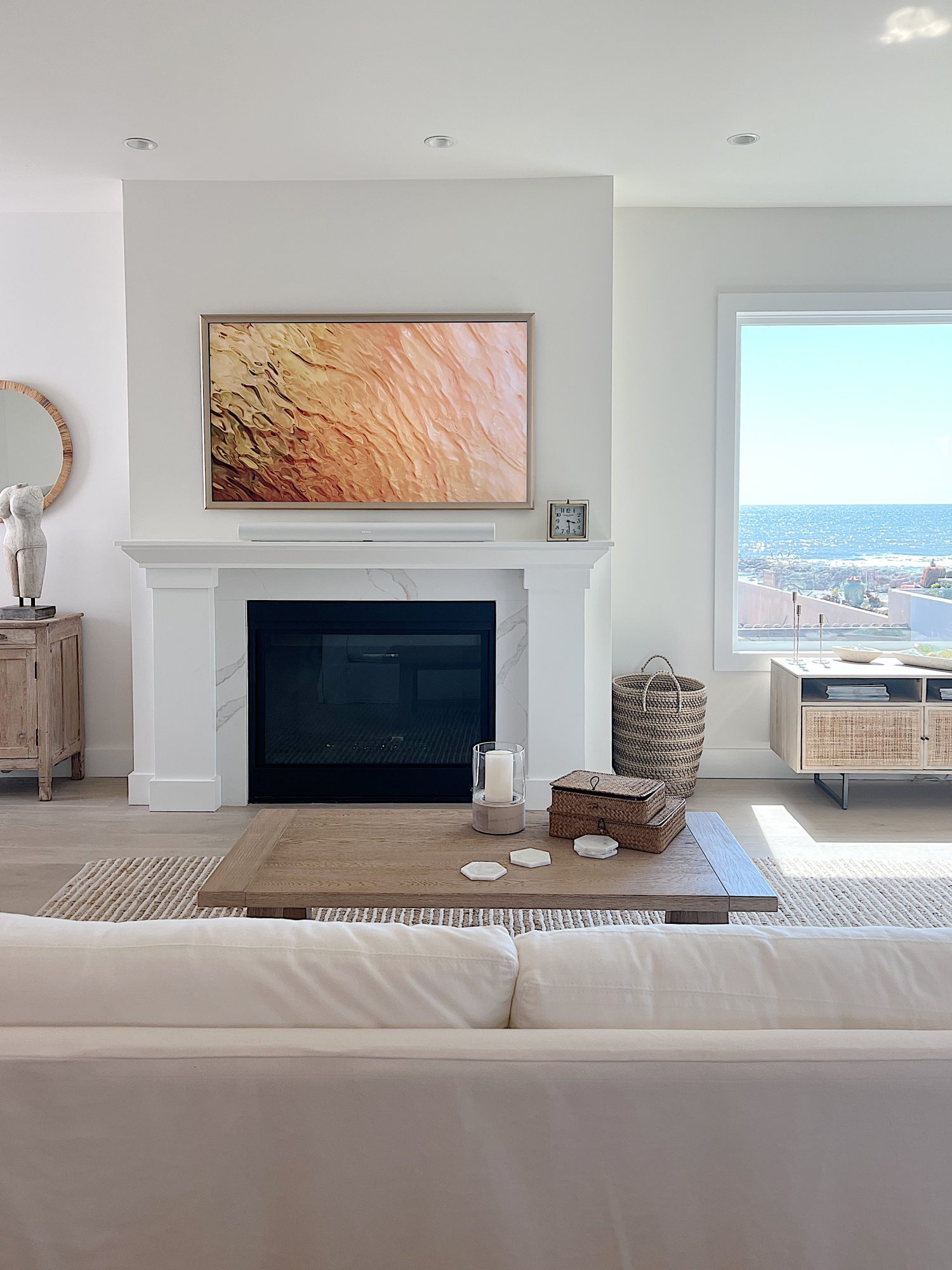 A modern living room with a white sofa, a fireplace with art above it, a large window showing an ocean view, and neutral-toned decor.