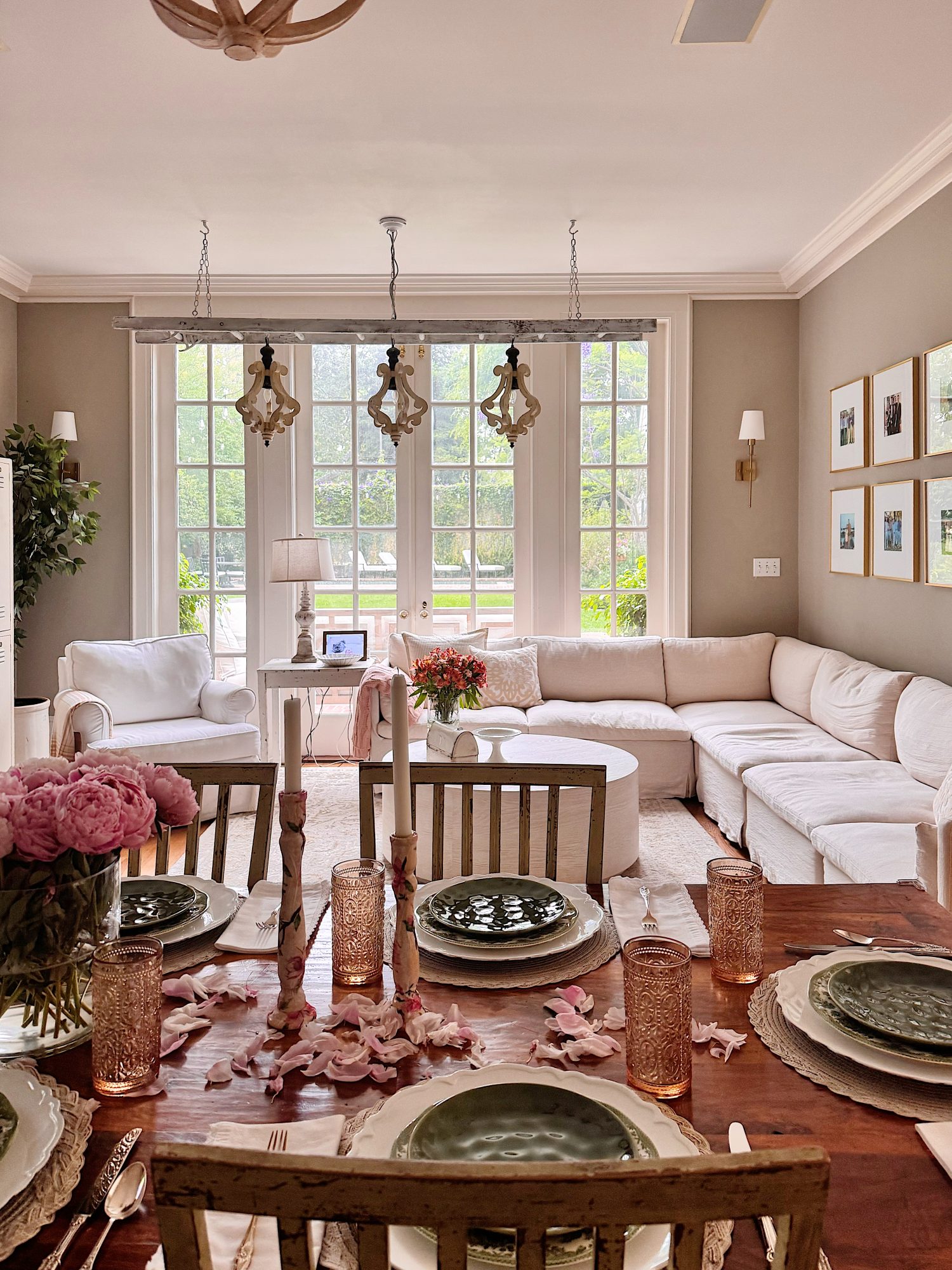 A bright, elegant living and dining room with a set table in the foreground, white sofa, armchairs, large windows, and soft natural light.