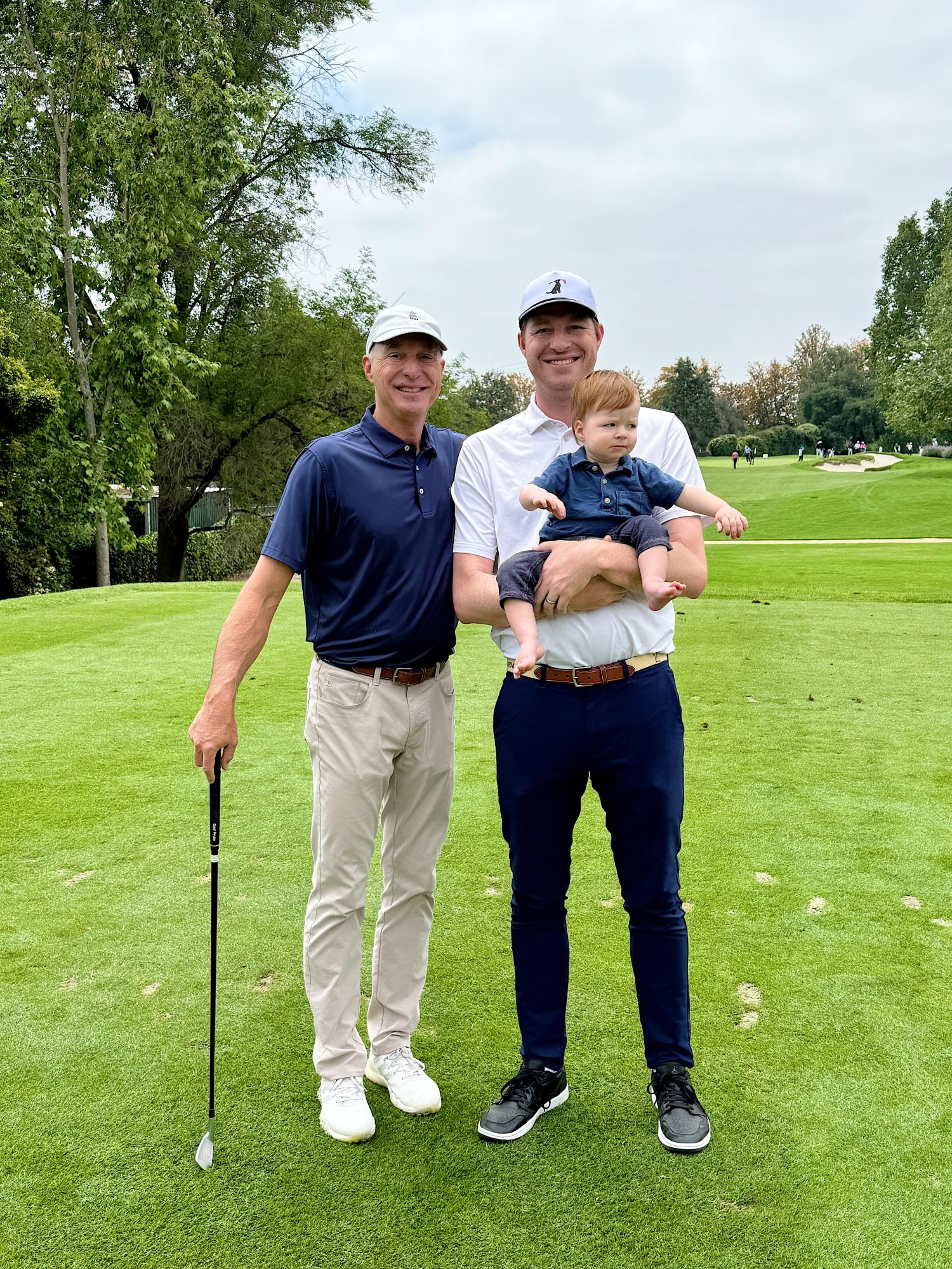 Two men stand on a golf course, one holding a putter and the other holding a baby. Trees and grass are visible in the background.
